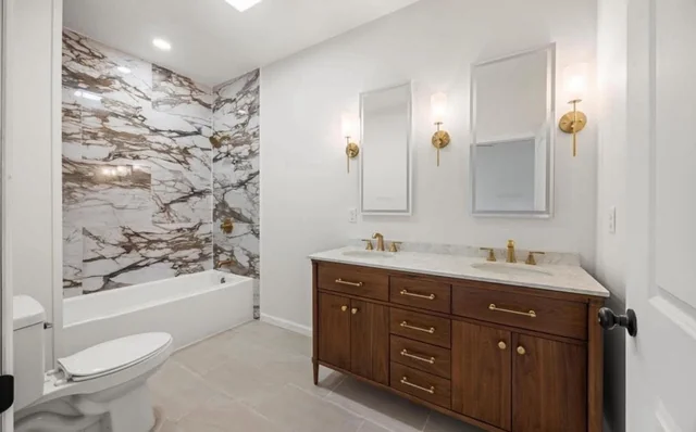 Brass sconces and recessed lights brighten a bathroom with wood cabinetry and marble tile